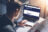 Closeup view of a man in eyeglasses working at sunny office on laptop while sitting at wooden table. The screen on his laptop is the OFNL account login page.